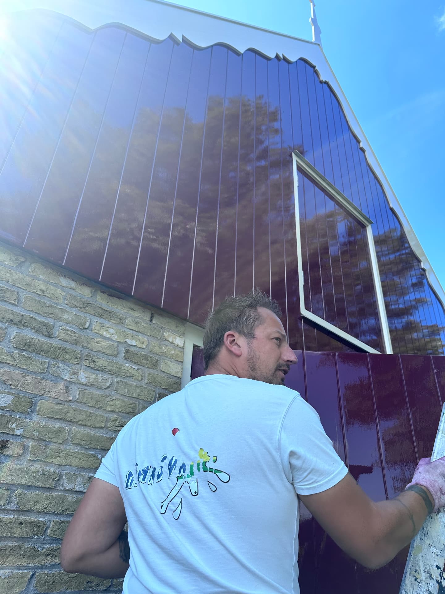 Schilder aan het werk op een ladder bij een glanzend bordeauxrode houten loodsgevel