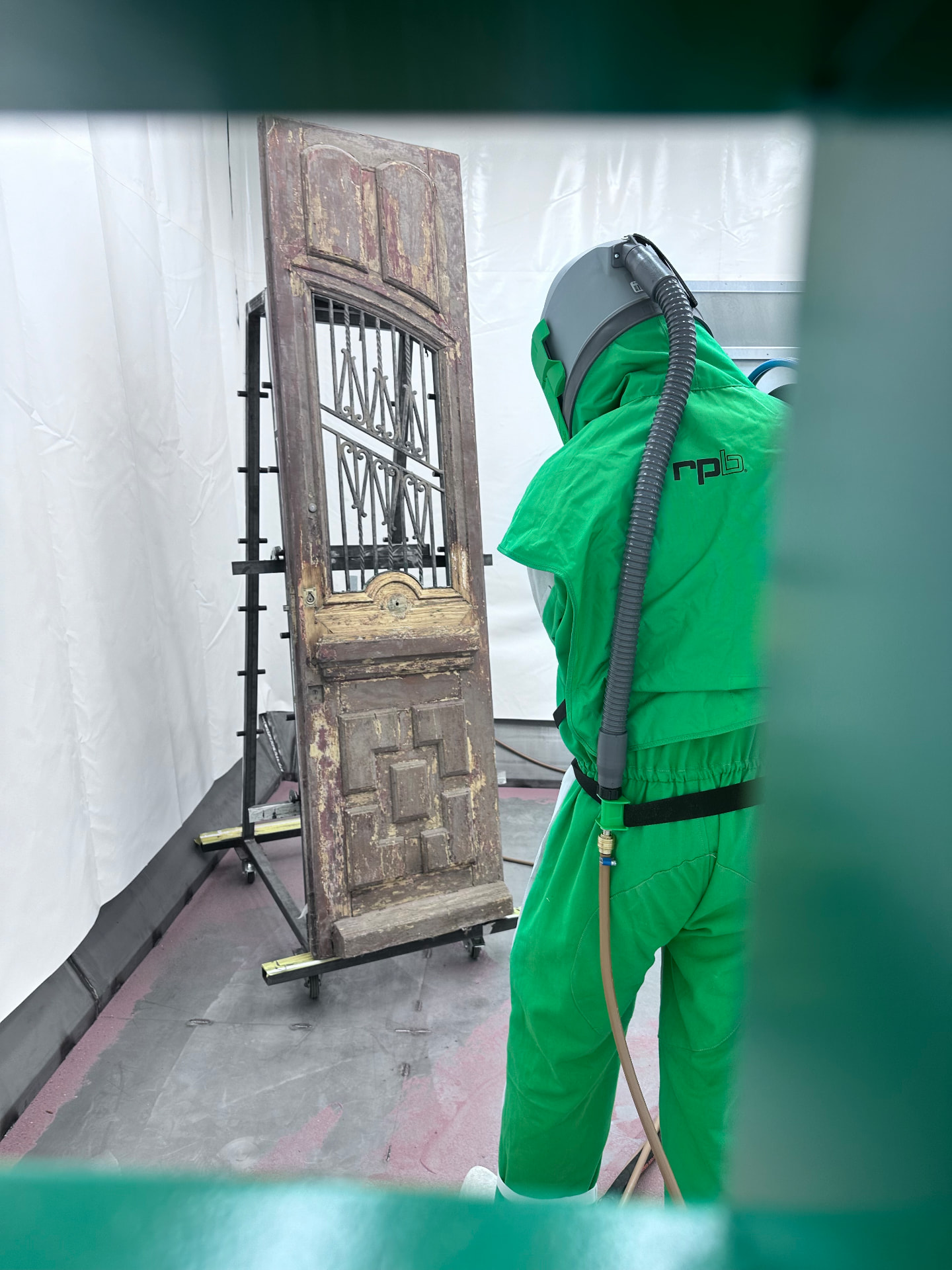 Vakman in beschermend straalpak die een verweerde houten deur met traliewerk straal in de cabine