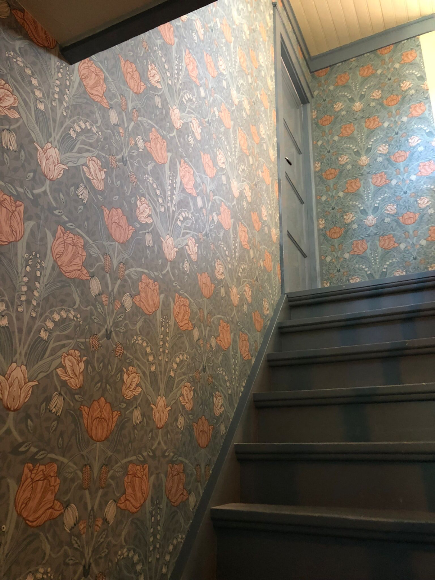 Traphal met donkerblauwe houten trap en een wand met bloemenbehang in zachte tinten